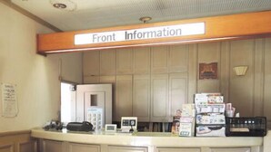 Un mostrador de recepción con un cartel que dice 'Front Information', un teléfono, una computadora y varios folletos informativos. Hay un cesto de basura y una alfombra de color oscuro en el suelo.