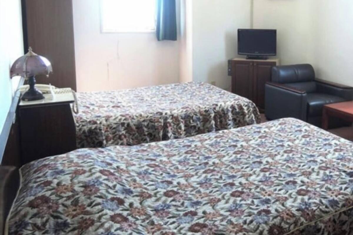 Habitación de hotel con dos camas, un escritorio con lámpara, televisión, una silla y una ventana con cortinas.