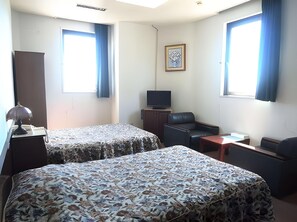 Ein Hotelzimmer mit zwei Betten, einem Schreibtisch mit Lampe, einem Fernseher, einem Sessel, einem kleinen Tisch und zwei mit Vorhängen versehenen Fenstern.