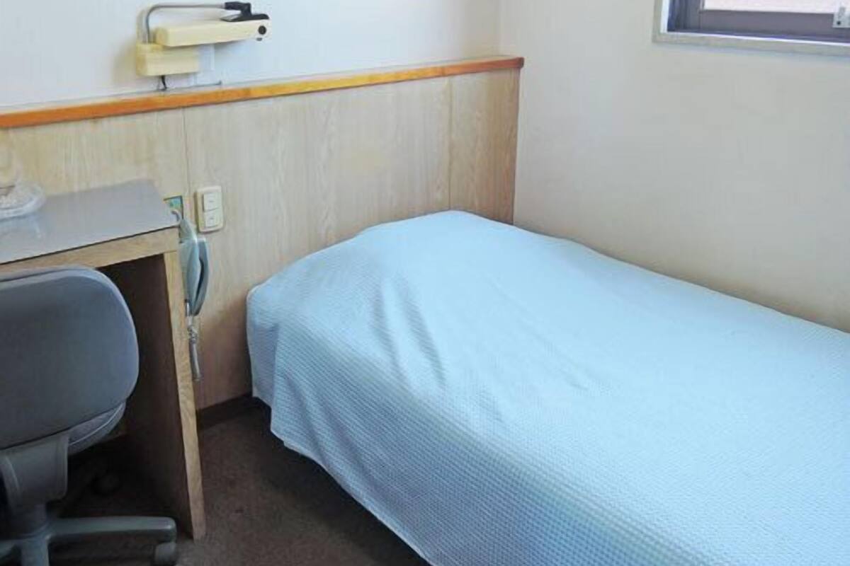 Una habitación pequeña con cama individual, escritorio y silla, ventana con persianas, y una lámpara fijada a la pared.