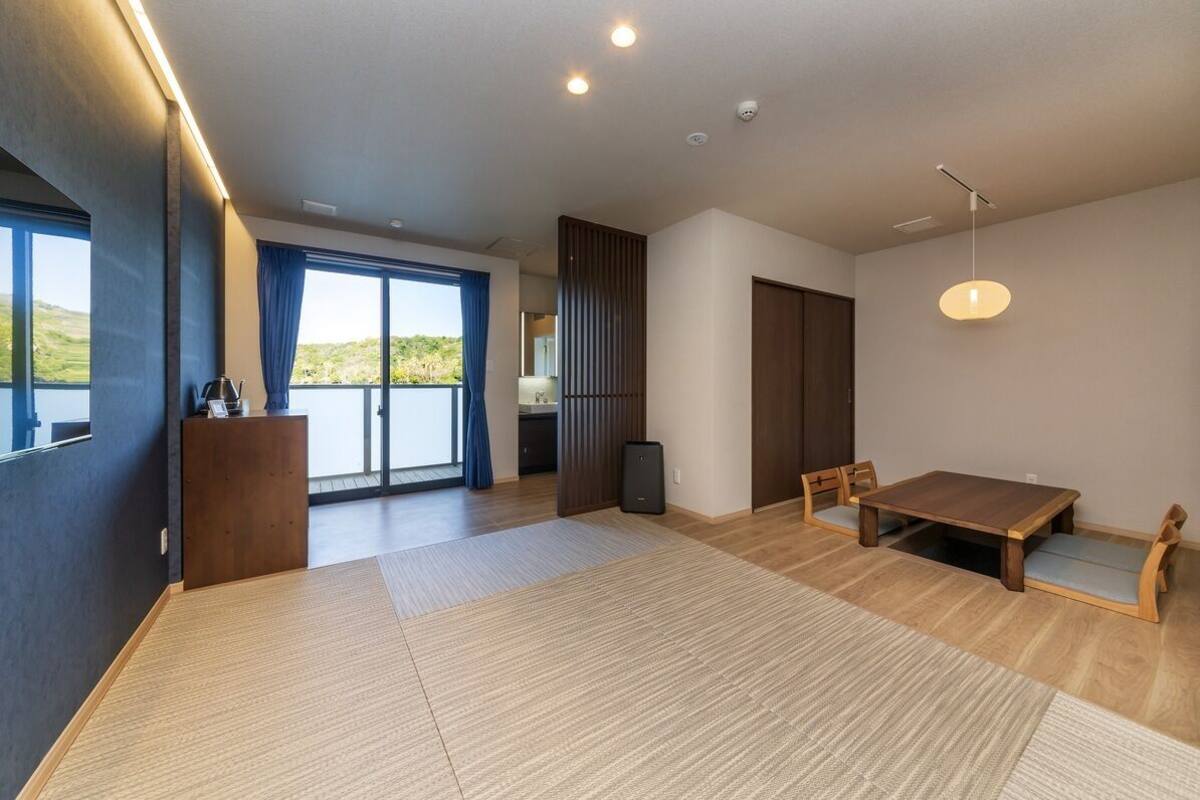 Ein geräumiger Raum mit einem großen Tisch, Stühlen, einer kleinen Kommode und einer Schiebetür zum Balkon.