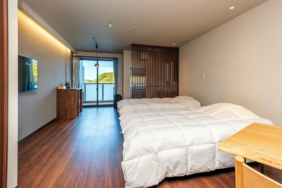 Ein Schlafzimmer mit einem großen Bett, Holzboden, einem Schreibtisch, Blick durch eine Schiebetür und einem Balkon.