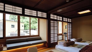 Una habitación japonesa tradicional con suelo de tatami, un banco de madera bajo, una cama con almohadas y una ventana con vistas a un edificio y árboles.