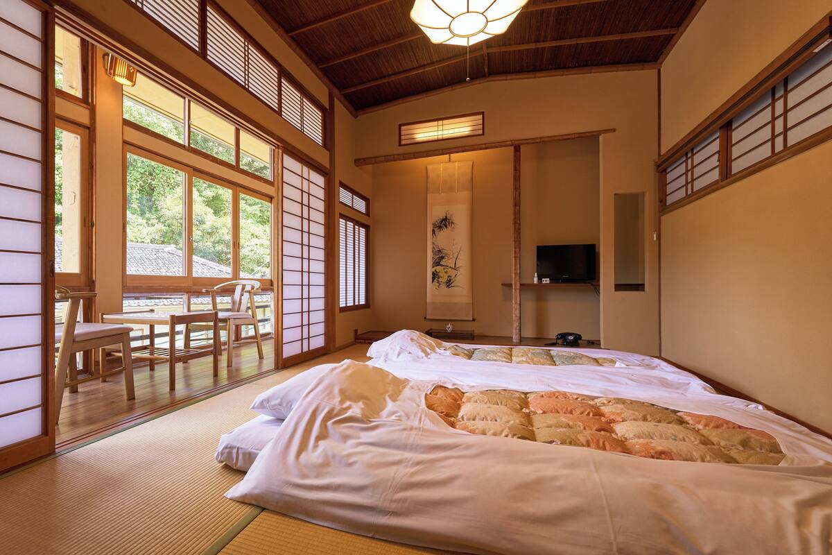 Ett traditionellt japanskt rum med tatamimattor på golvet, en säng med ett täcke, en tv och en skjutdörr som leder ut till ett område utomhus.