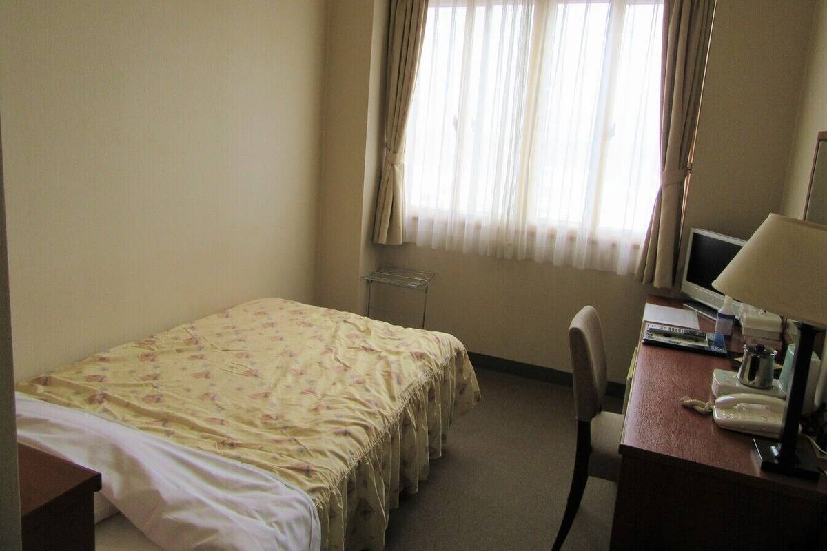 Ein Hotelzimmer mit Bett, Schreibtisch mit Computer, Stuhl und Fenster mit Vorhängen.