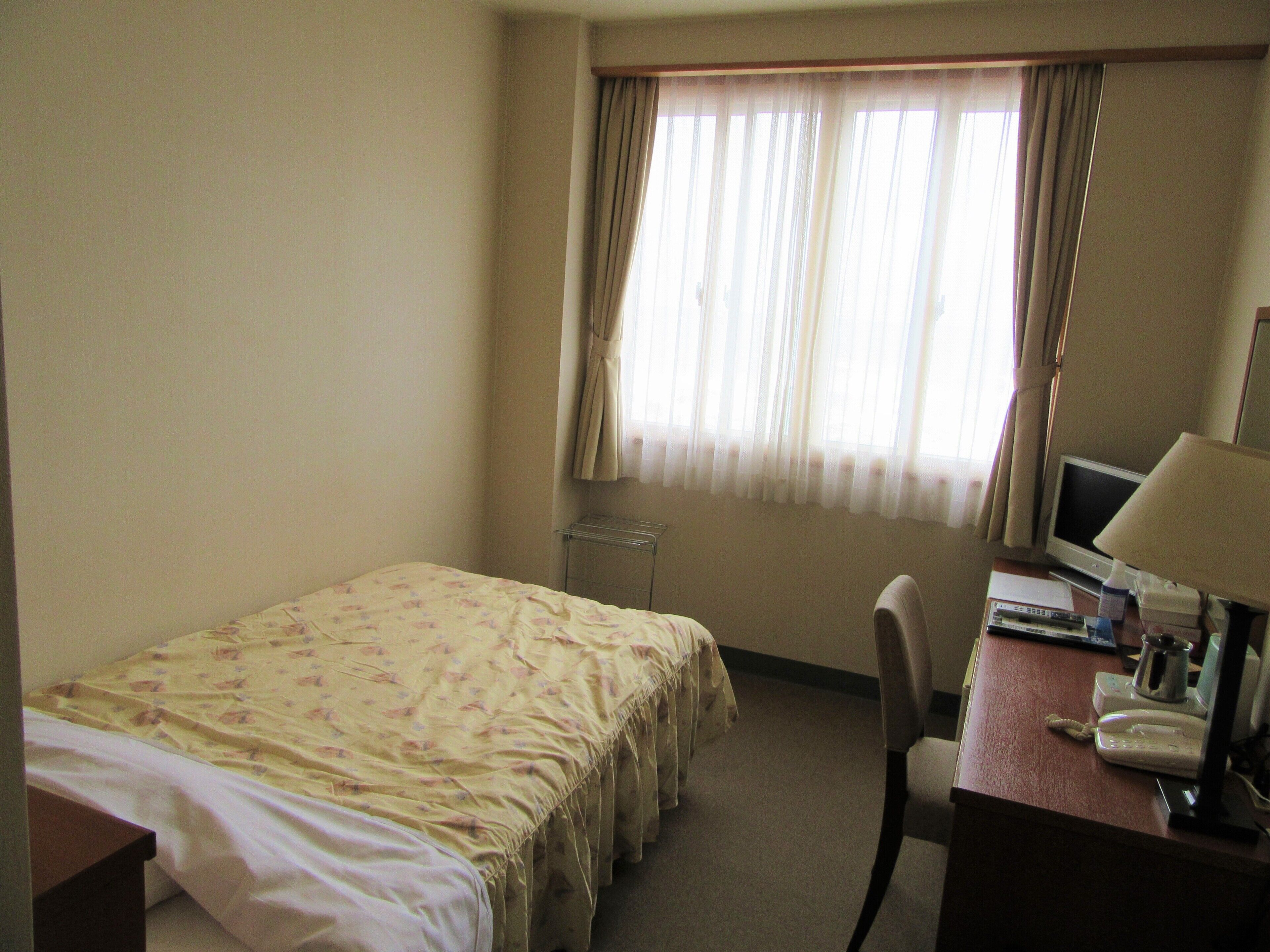 A hotel room with a bed, a desk with a computer and lamp, a chair, and a window with curtains.