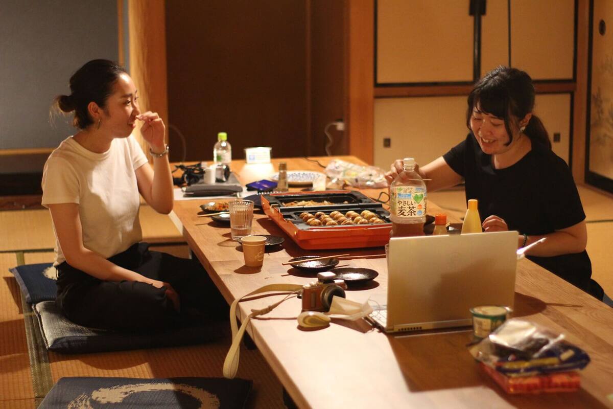 Two people sitting at a wooden table with a laptop, a tray of food, and various items like a water bottle and a glass.