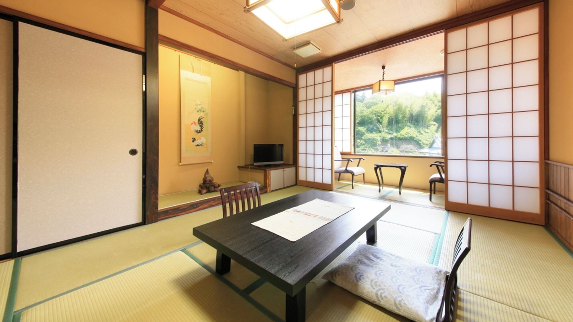 Perinteinen japanilainen huone, jossa on tatamimatto, matala pöytä, tuoleja, televisio ja näkymä vehreään maisemaan.