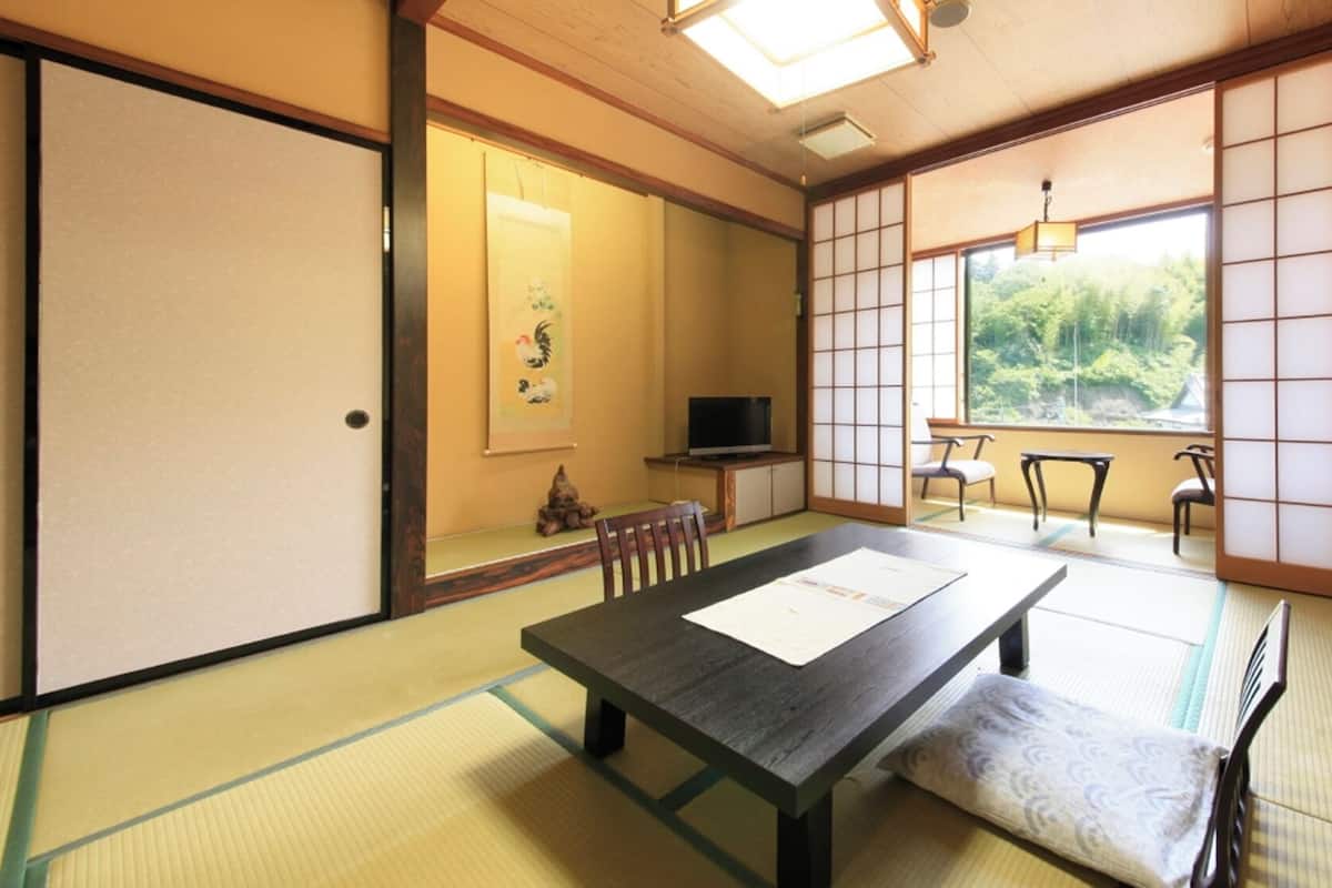 A traditional Japanese room with tatami flooring, a low table, chairs, a television, and a view of greenery.