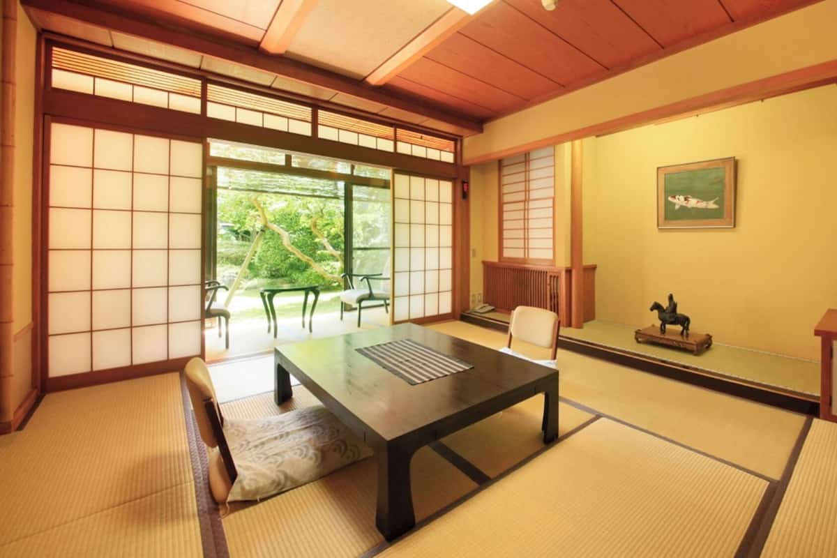 A traditional Japanese room with tatami flooring, a low table, a chair, a cabinet, and a window with a view of greenery.