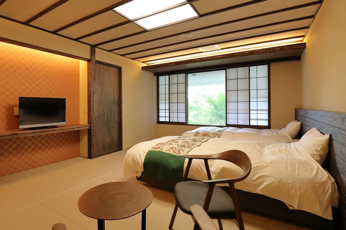A hotel room with a bed, a TV on a stand, a chair, a small table, and a window with a view of greenery.