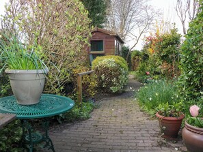 Un sentier de jardin mène à une structure en bois, qui est entourée de différentes plantes et d’une table en métal vert.