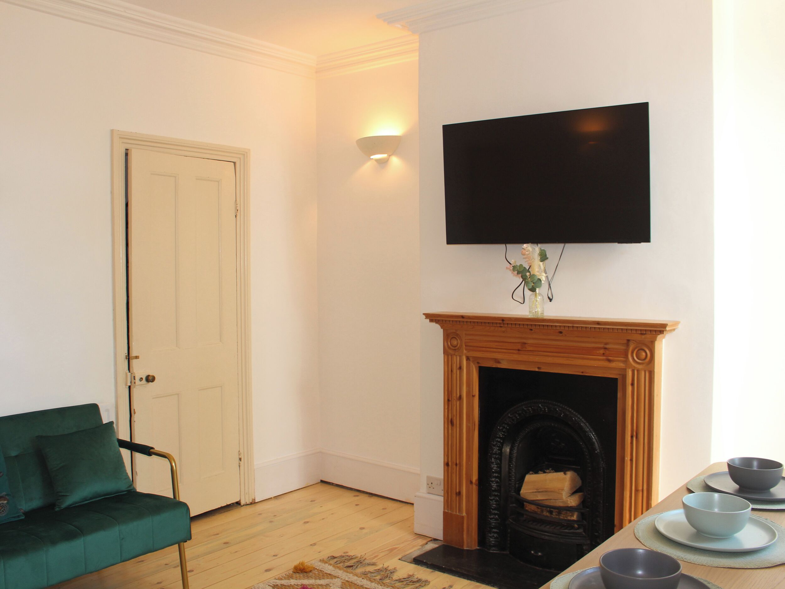 Una habitación con un sofá verde, una chimenea, un televisor de pantalla plana montado encima y una mesa de comedor con cuencos.