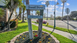 Placa para aluguel de temporada Sunset Shores, com vista clara da vegetação ao redor e uma rua ao fundo.