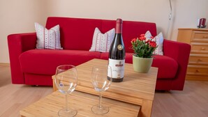 Un sofá rojo, una mesa de madera con una botella de vino y copas, y una planta en maceta con flores rojas.