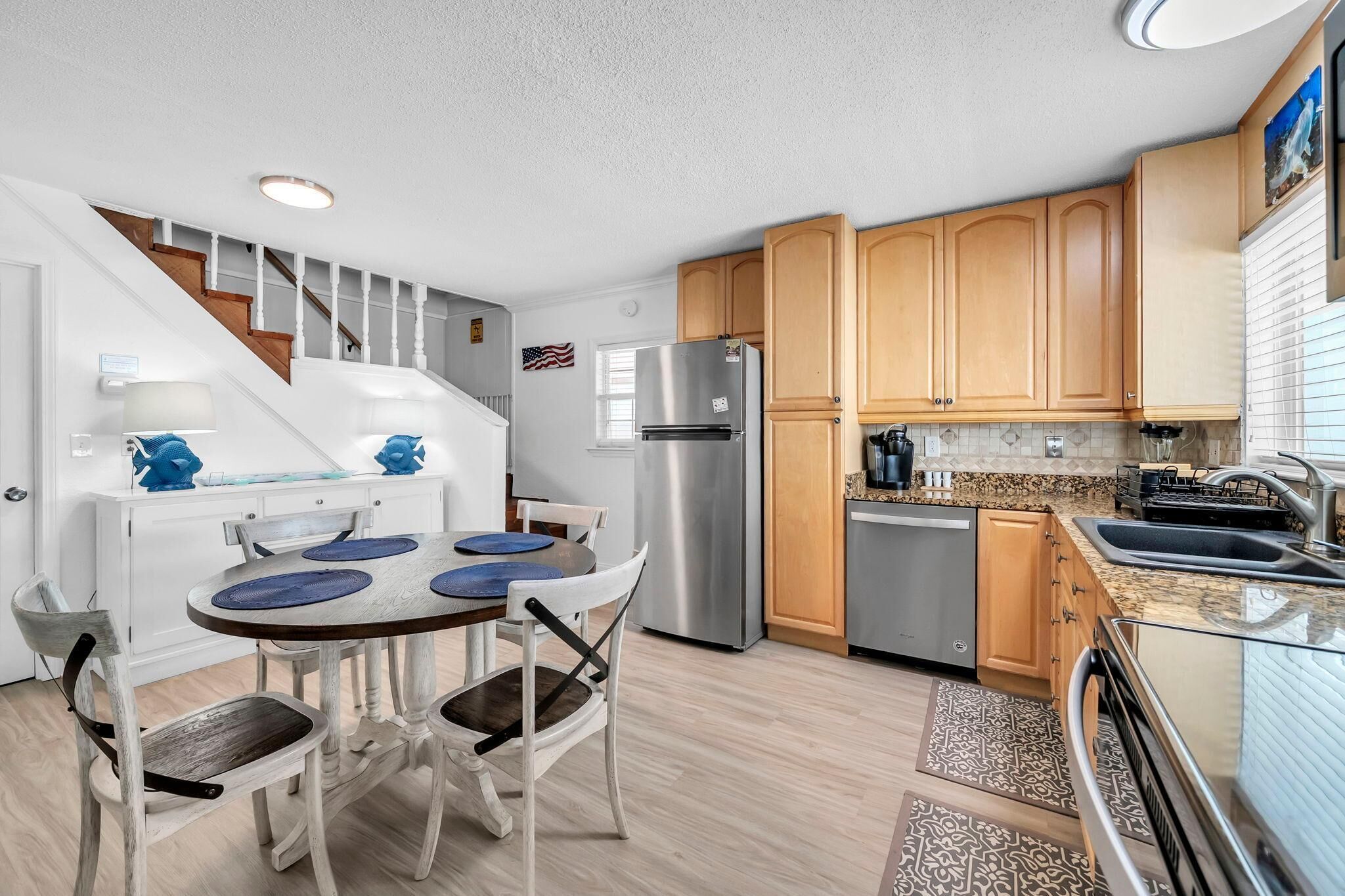 A modern kitchen with a stainless steel refrigerator, wooden cabinets, a dining table with blue placemats, and a staircase leading to another level.