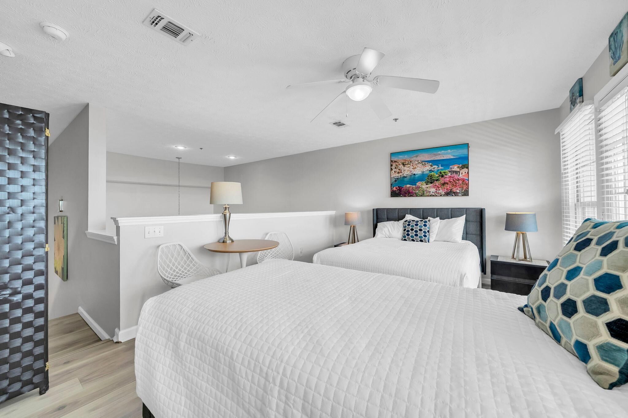 A hotel room with two beds, a ceiling fan, a painting, and a window with blinds.