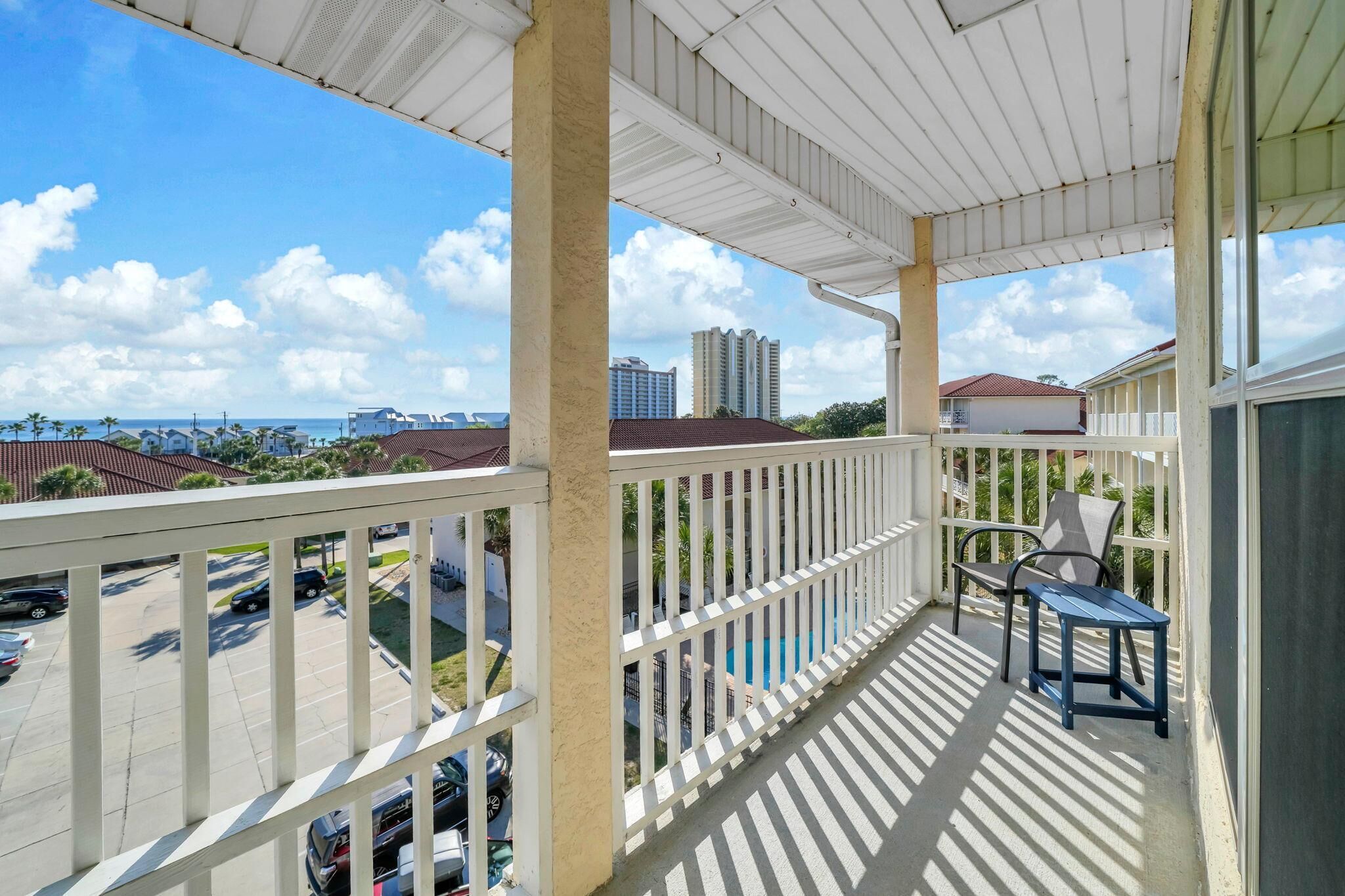 A balcony with a view of a parking lot and buildings, a pool, and a covered area with chairs.