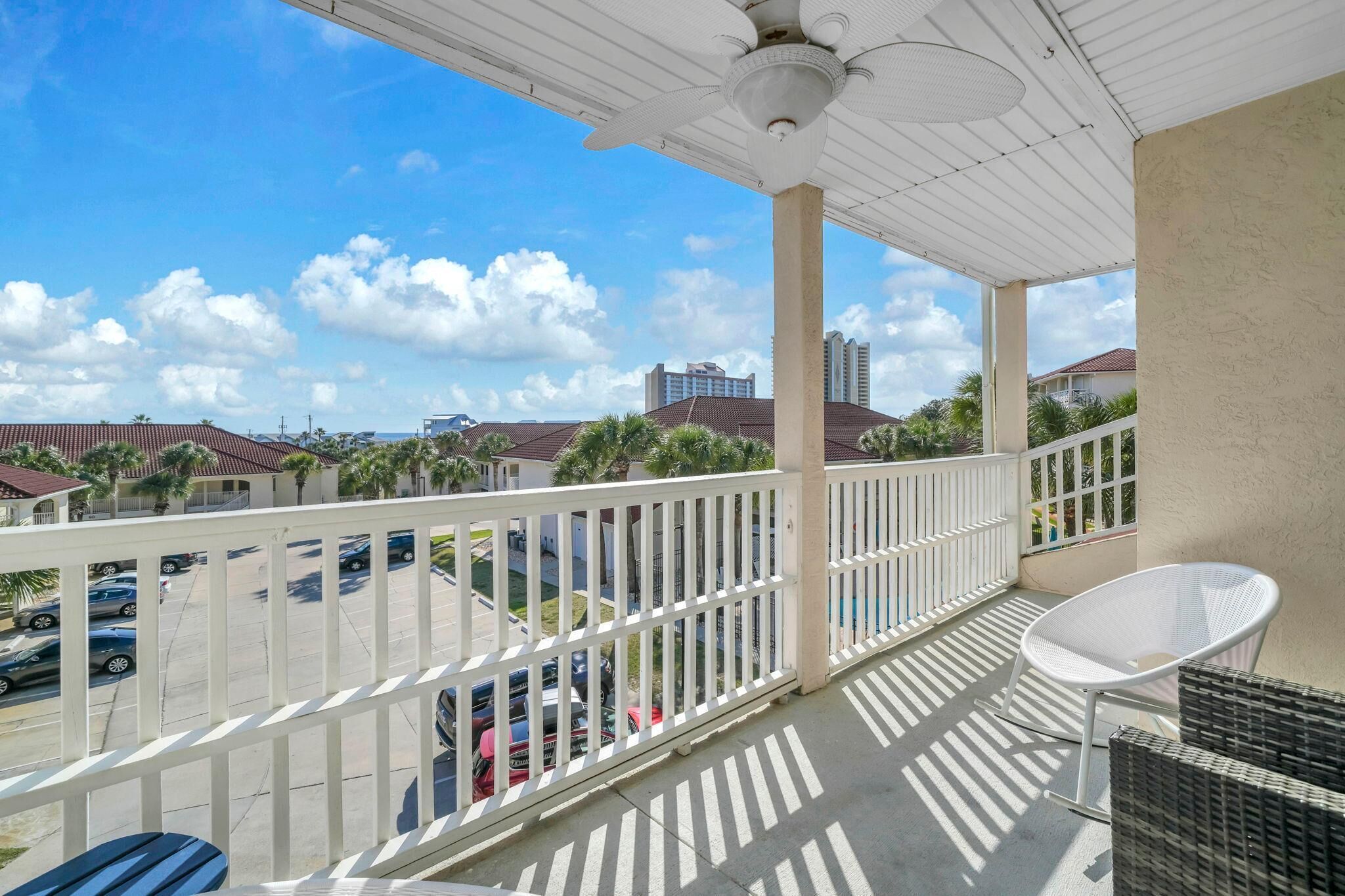 A balcony with a white railing, a ceiling fan, and a view of a parking lot and buildings.