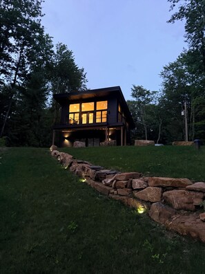 Une cabane en bois à deux étages, dotée de grandes fenêtres et d’un chemin de pierre menant à l’entrée.