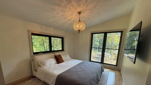 Quarto com cama, tapete, lustre e duas janelas com vista para uma floresta.