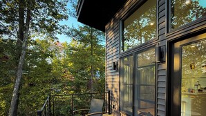 Um deck de madeira com área de estar externa, guarda-corpo e vista para uma área arborizada.