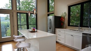Cozinha moderna com ilha central, banquetas, geladeira e uma janela ampla com vista para árvores.