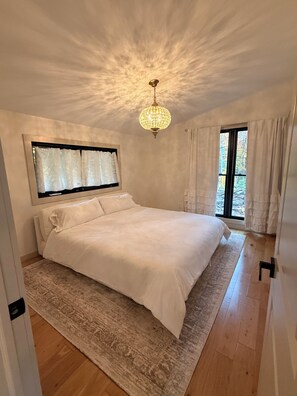 Ein Schlafzimmer mit einem großen Bett, einer Kristallleuchtre, einem Fenster mit Vorhängen und Blick auf Bäume.