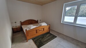 Quarto com cama de madeira, mesa de cabeceira, tapete verde, janela com vista para árvores e teto de madeira.