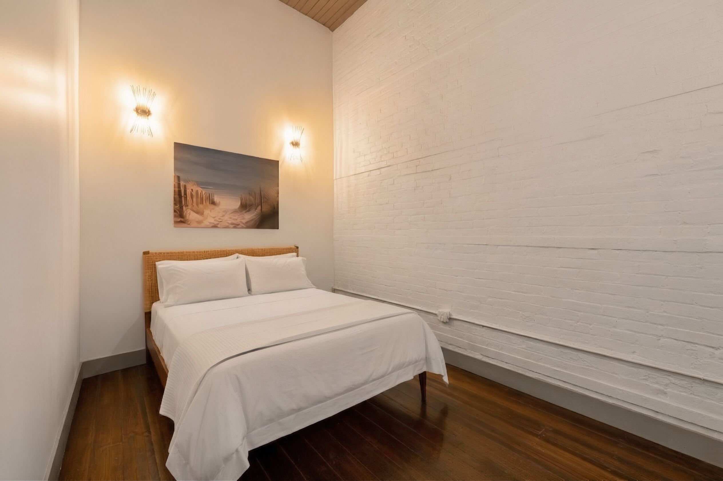 Un dormitorio con cama de madera, ropa de cama blanca, un cuadro colgado en la pared y dos apliques.