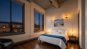 A bedroom with a large bed, two wall sconces, a painting, and a view of the city at dusk.