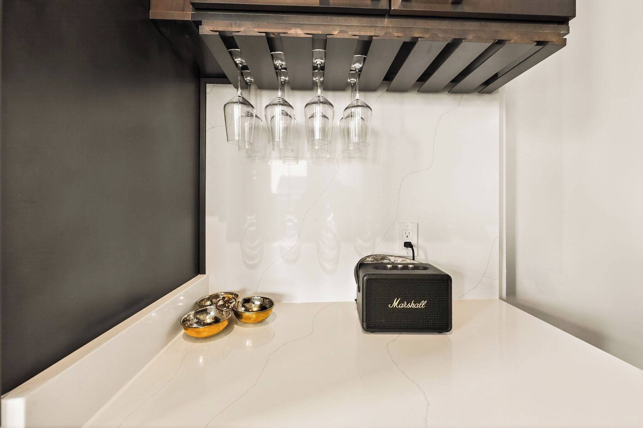 A kitchen with a marble countertop, hanging wine glasses, a black speaker, and two pet bowls.