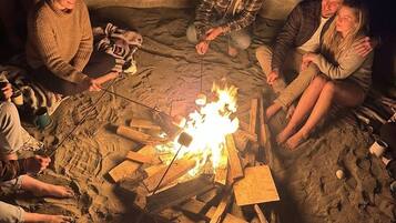 A group of people around a campfire in a sandy outdoor setting.