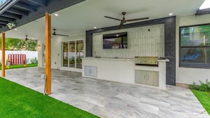 Ein möblierter Außenbereich mit Grill, Deckenventilatoren und einem an der Wand montierten Fernseher.