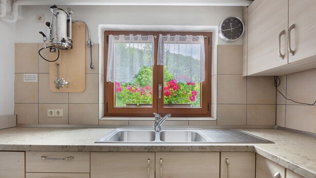 A kitchen with a double sink, wooden cabinets, a window with lace curtains, and a clock on the wall.