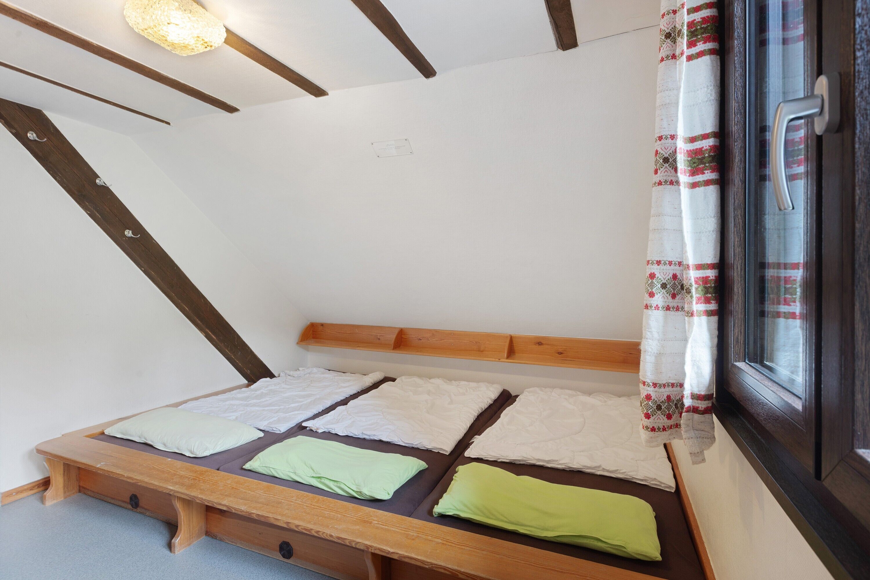 A small, single-bed room with a sloped ceiling, wooden beams, a window with a curtain, and a built-in bench at the foot of the bed.