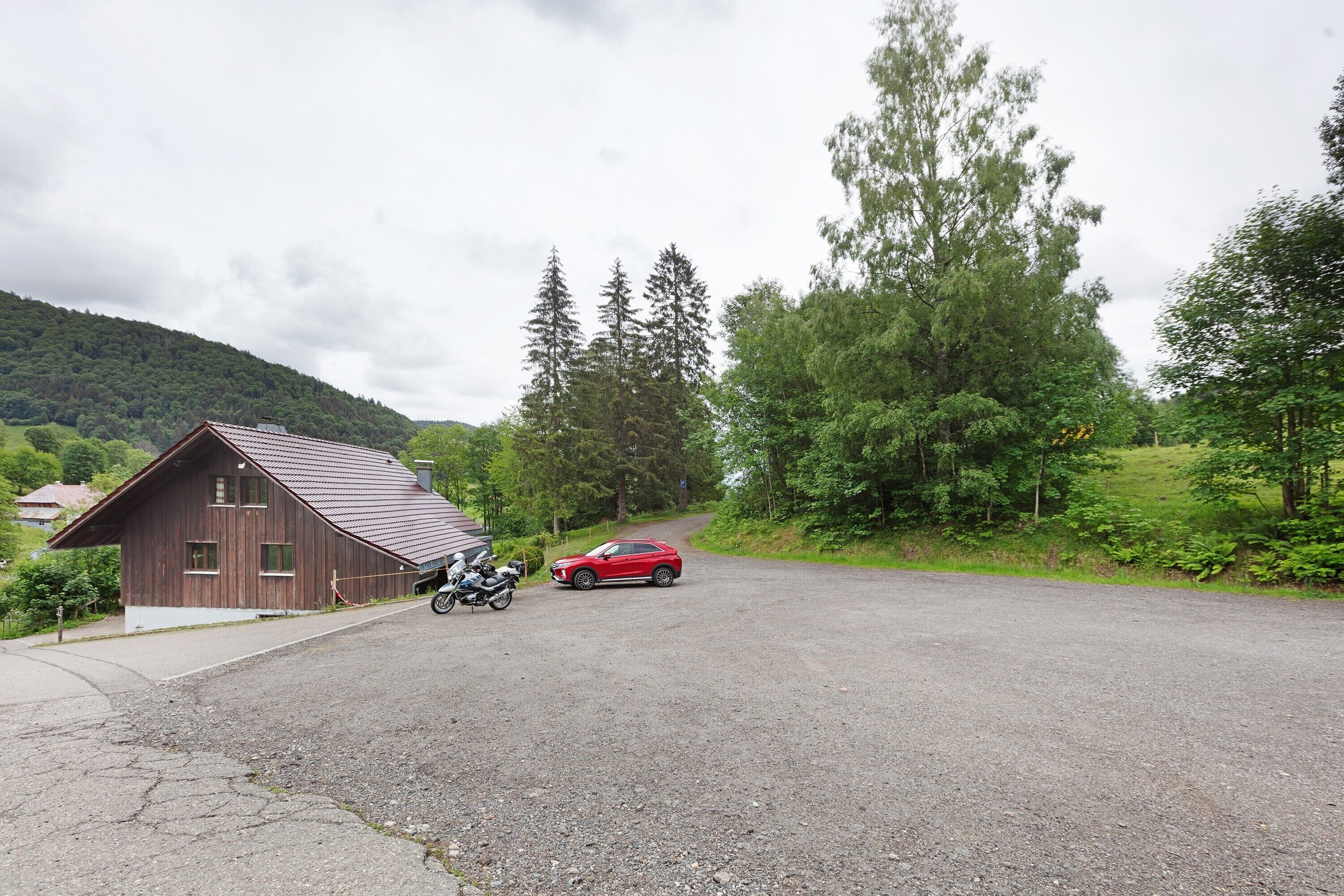 Uma estrada rural com um edifício de madeira, um carro vermelho e uma motocicleta estacionada.