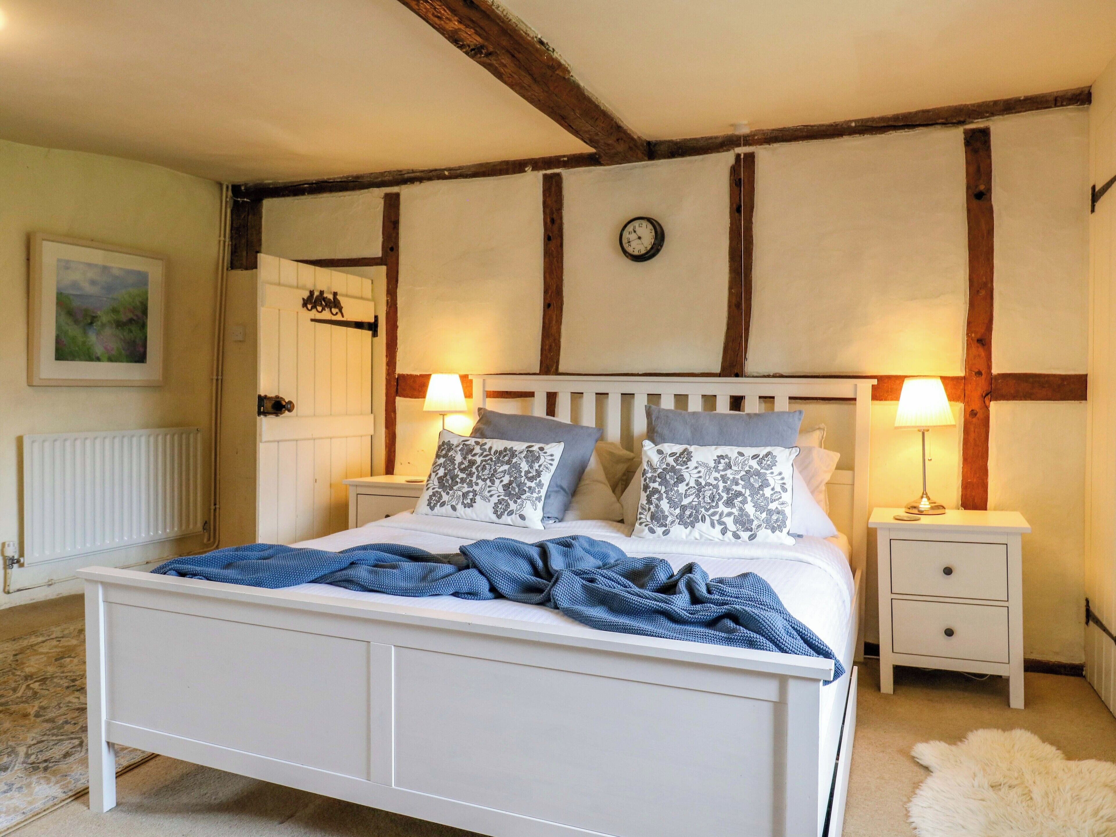 A bedroom with a white bed, two bedside tables with lamps, a wooden ceiling, and a clock on the wall.