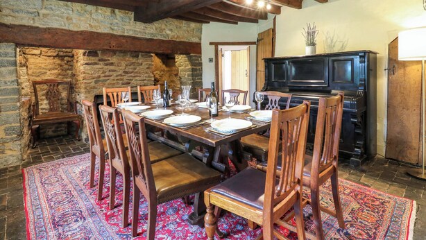 A dining area with a stone wall, wooden beams, a piano, and a dining table set for six. - Manor Farm Cottages (Milton Keynes)