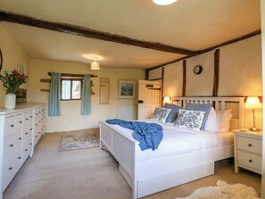 A bedroom with a bed, bedside tables, a dresser, a window with curtains, a radiator, and a clock on the wall. - Manor Farm Cottages (Milton Keynes)