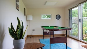 A room with a pool table, a potted plant, a lamp, a dartboard, and a window with a view of greenery. - Manor Farm Cottages (Milton Keynes)