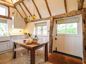 A rustic kitchen with wooden beams, a dining table with chairs, a window with a view, and a door. - Manor Farm Cottages (Milton Keynes)