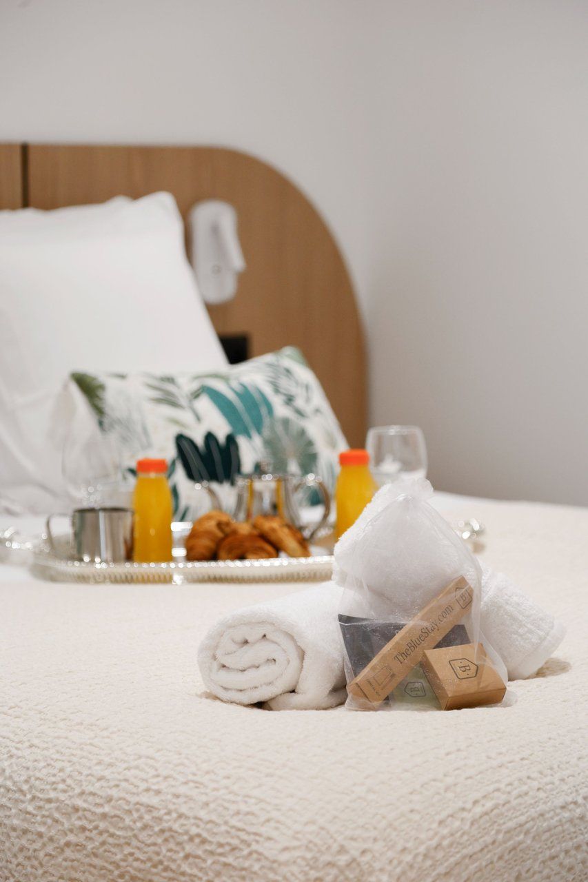 A neatly made bed with a rolled white towel, a tray with croissants and orange juice, and a plastic bag with a logo.
