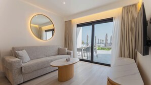 Une chambre d’hôtel moderne équipée d’un canapé, d’une table basse ronde, d’un téléviseur à écran plat et d’une porte-fenêtre coulissante donnant sur un balcon aménagé avec du mobilier d’extérieur.