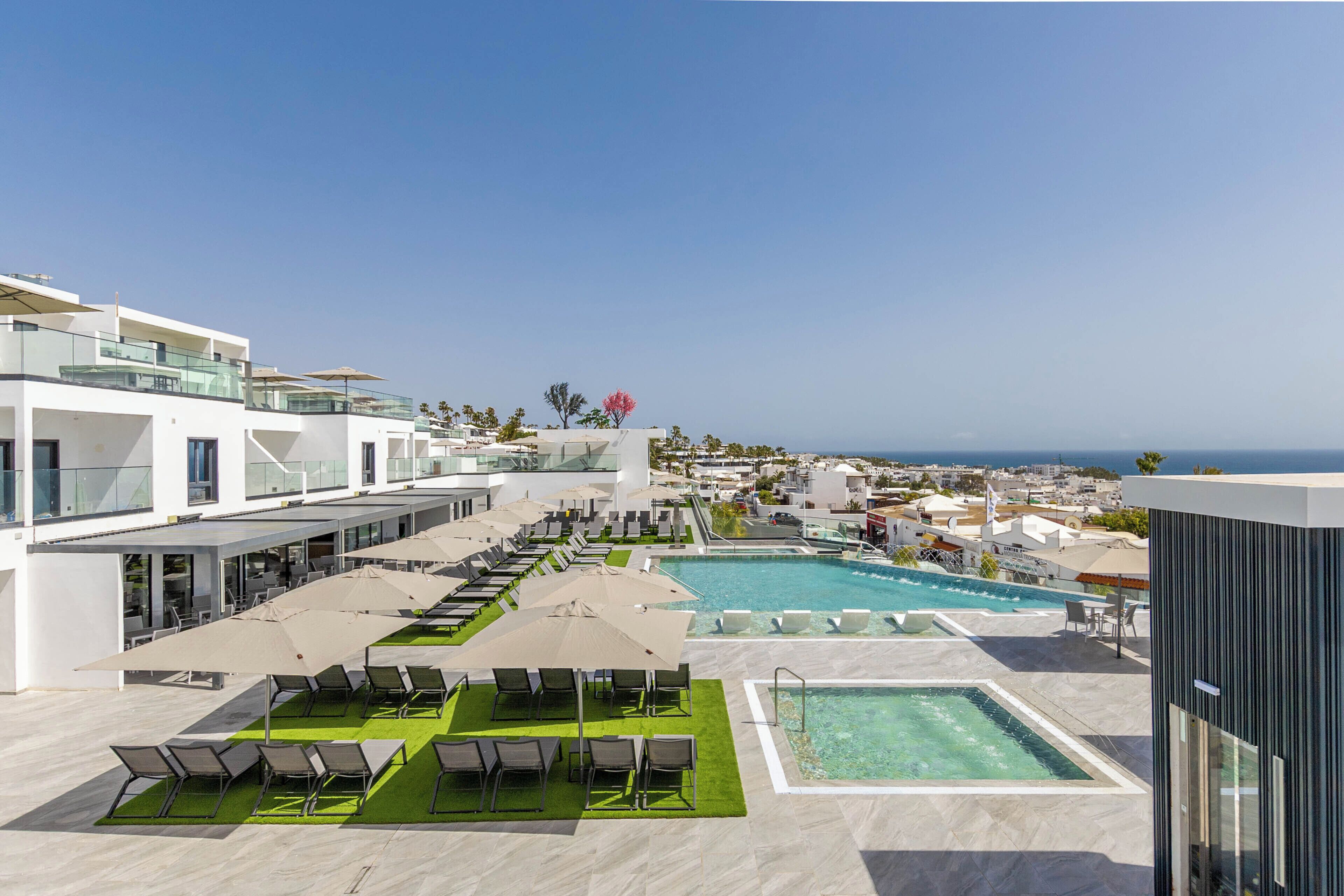 A rooftop pool area with sun loungers and umbrellas, a clear view of the ocean, and modern architecture.