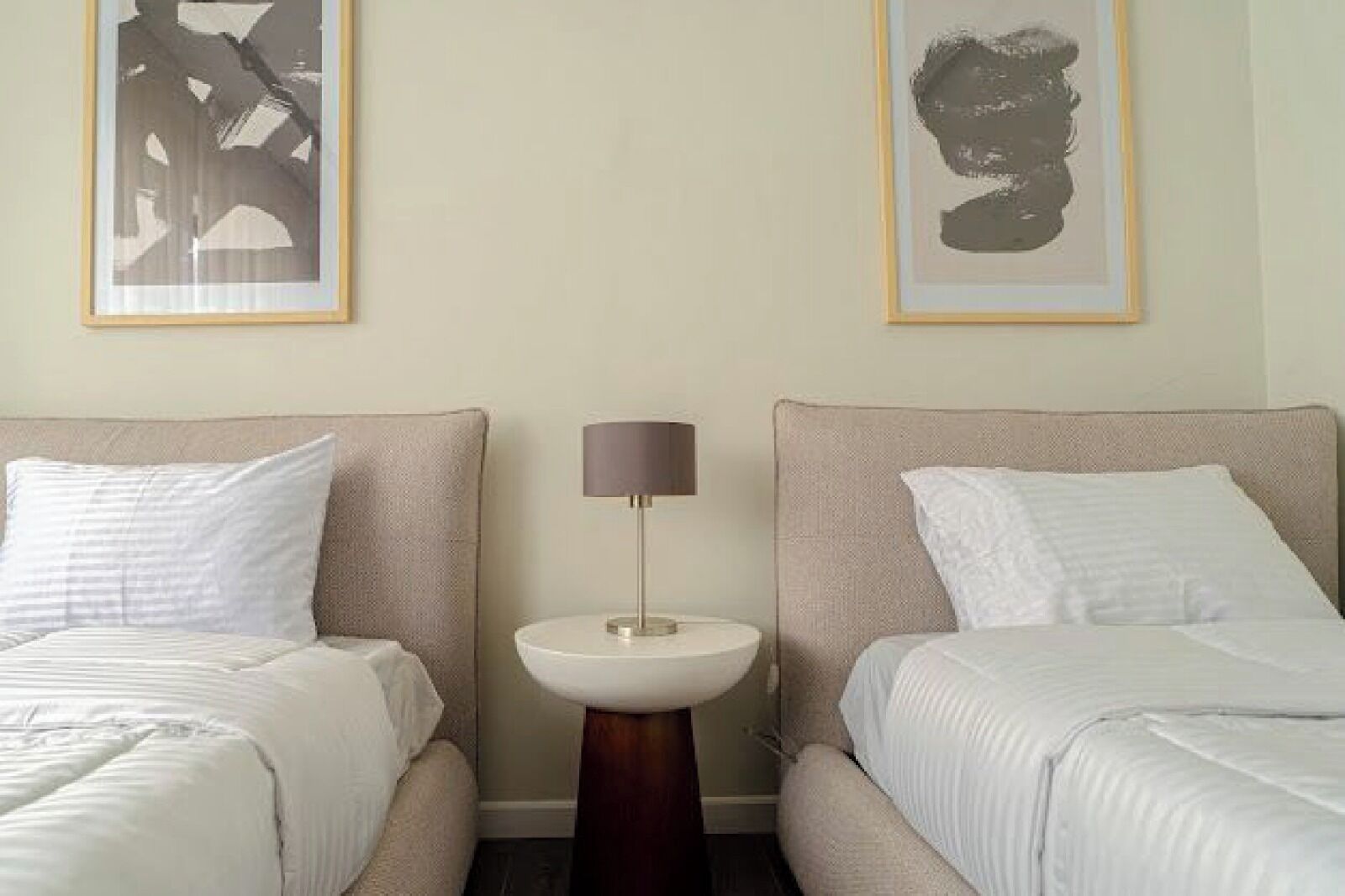 A hotel room with two beds, a round bedside table with a lamp, and two framed abstract paintings on the wall.