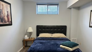 A bedroom with a dark blue bedspread, a headboard, a bedside lamp, a small table with a tissue box, and a window with blinds.