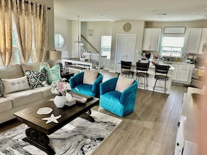 A living room with a sofa, two blue armchairs, a coffee table with decorative items, a dining area with a table and chairs, a kitchen with white cabinets, and a staircase leading to another level.