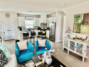 A modern kitchen with white cabinets, a stainless steel refrigerator, and a microwave. There is a dining area with a blue sofa and a coffee table.