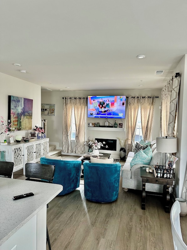 A modern living room with a flat-screen TV, blue upholstered chairs, a white sofa, and a dining area with a table and chairs.